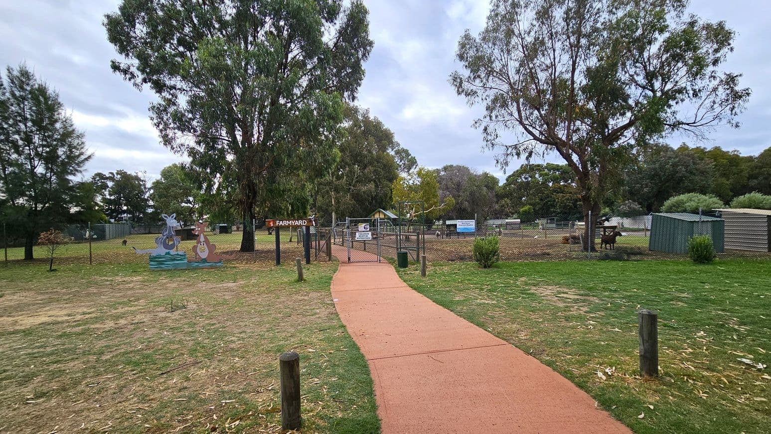A red pathway leads to a fenced area labelled "Farmyard" in the centre. On the left, there are cut-out figures of kangaroos near a tree. To the right, a green shed and a fenced area with animals are visible. The background is filled with tall trees and greenery. The ground is a mix of grass and dirt, with some fallen leaves scattered around.