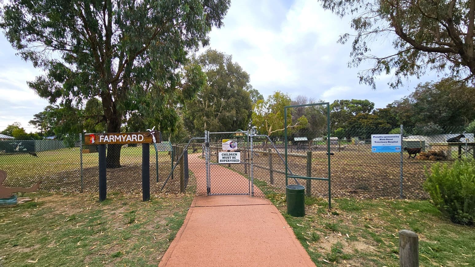 A farmyard entrance with a chain-link gate and a sign reading "CHILDREN MUST BE SUPERVISED". To the left, a wooden sign says "FARMYARD" with decorative elements. The ground is a mix of grass and a reddish path leading to the gate. Behind the gate, there are trees and some farm structures. A green bin is near the entrance. The area is enclosed by a fence, and there are signs on the right side of the gate.