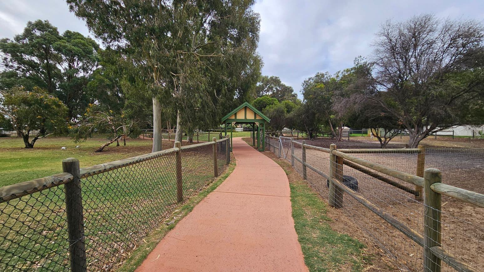 A red paved path leads through a park, flanked by wooden fences on both sides. On the left, there's a grassy area with tall trees and some yellow flowers. On the right, the ground is more barren with sparse trees. In the centre, a small green-roofed shelter stands over the path. The background is filled with more trees, creating a lush, natural setting.