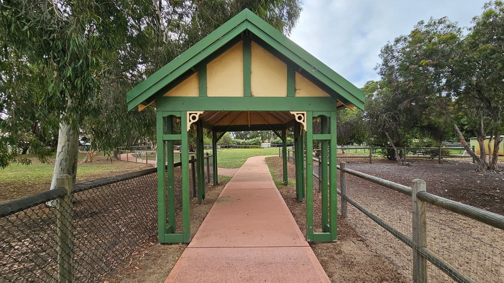 A wooden gazebo with a green frame and beige panels stands over a paved path. The path extends through the gazebo and curves into the distance. On either side, there are wire mesh fences supported by wooden posts. Trees with dense foliage surround the area, providing shade. The ground is covered with a mix of grass and dirt.