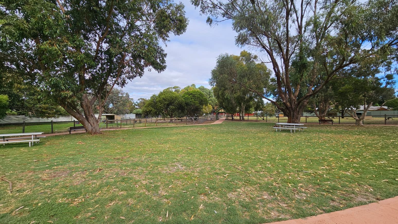 A grassy park area with scattered fallen leaves. On the left, a large tree stands near a white picnic table. In the centre, a path leads to a fenced area with more trees and a small structure in the distance. On the right, another large tree is near a second picnic table. The background features a fence and additional trees, creating a natural, open space.