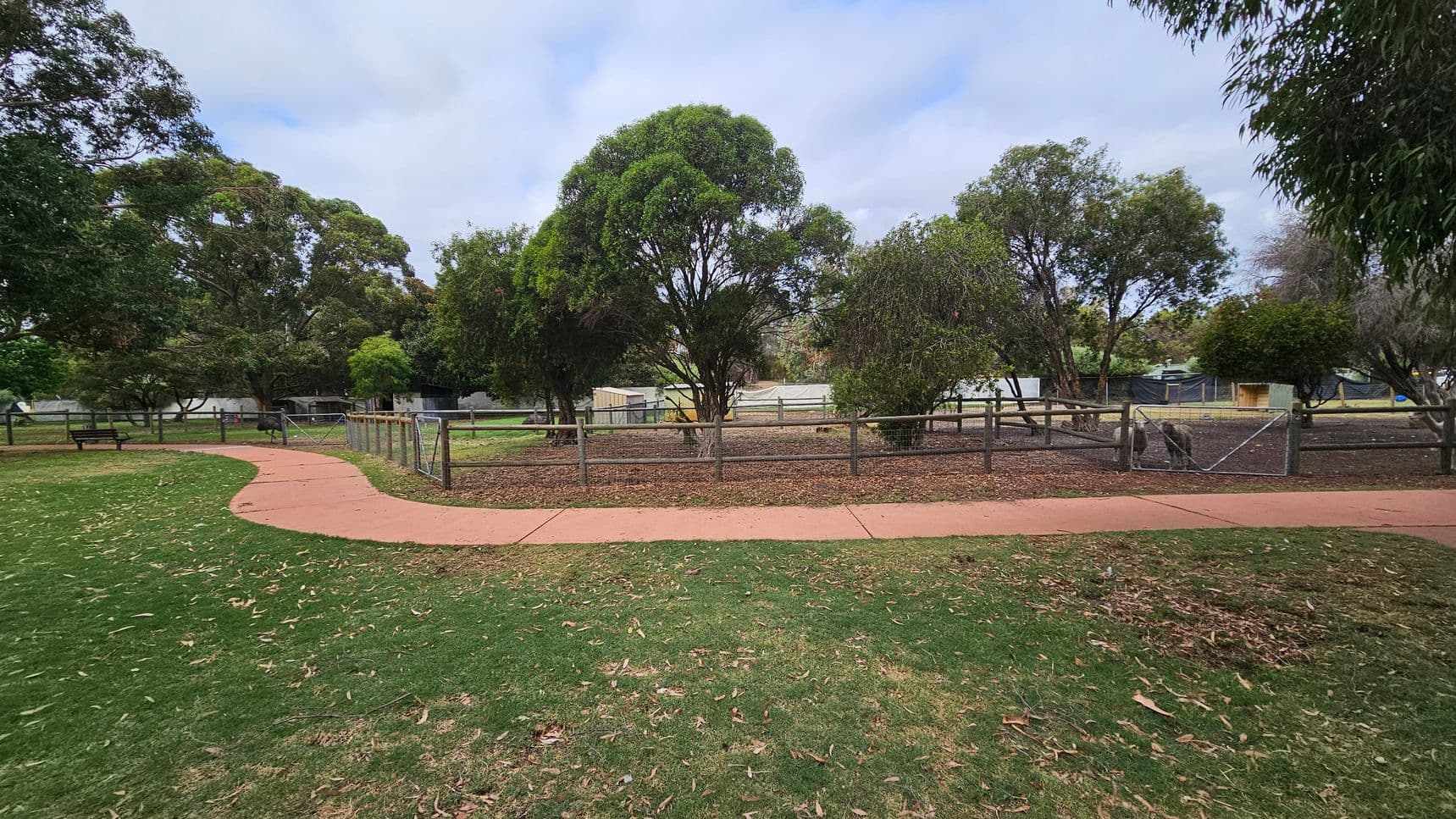 A park scene with a winding red path on the left, bordered by green grass and scattered leaves. In the middle, a fenced area contains several trees with dense foliage. The background features more trees and some structures, possibly sheds or small buildings. A bench is visible on the left side near the path. The overall setting is open and natural, with a mix of greenery and man-made elements.
