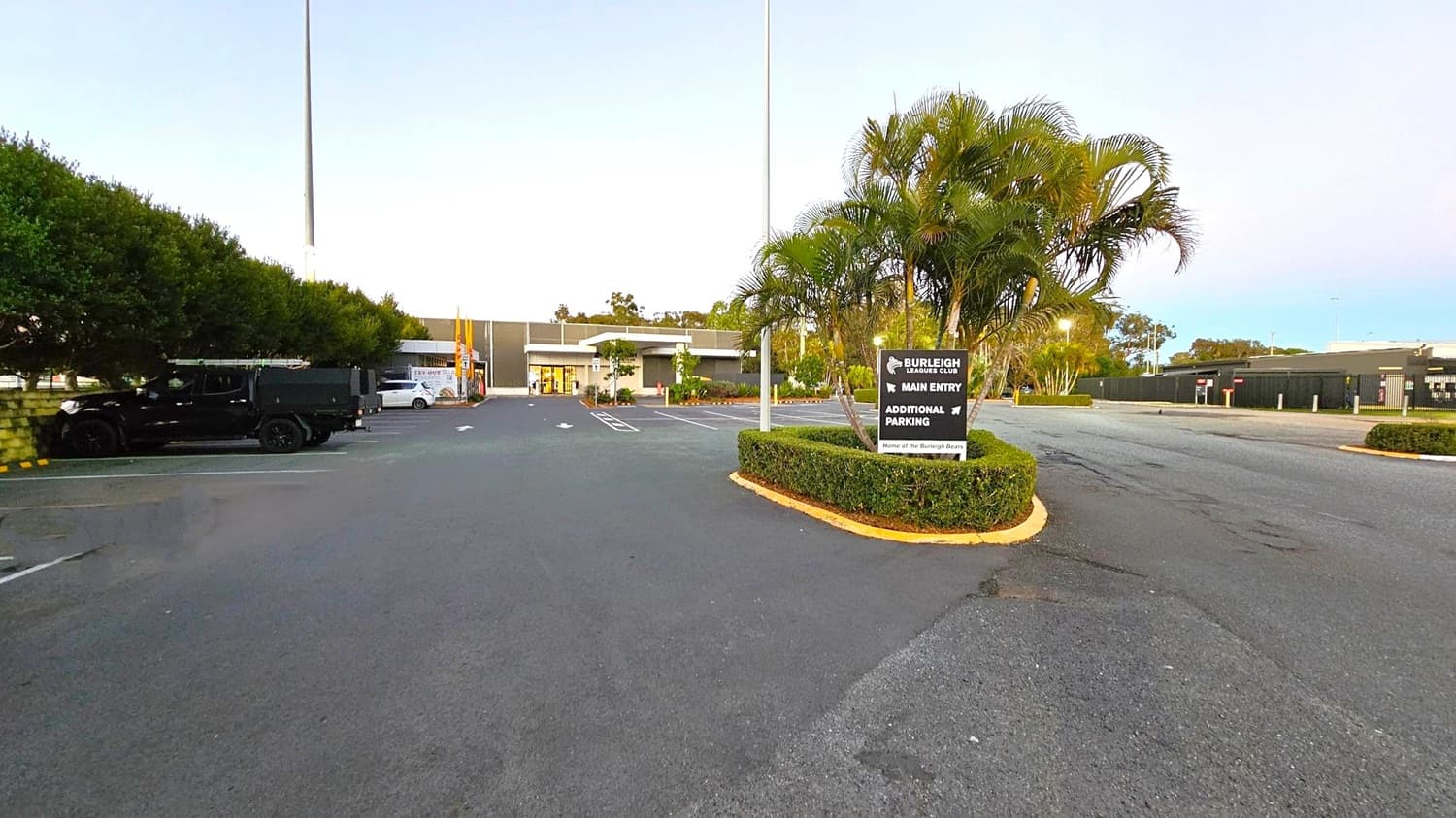 A car park with a sign for the Burleigh Leagues Club is in the foreground, surrounded by neatly trimmed bushes and palm trees. On the left, there are parked vehicles, including a black truck and a white car, next to a row of trees. The main building is in the centre, with a visible entrance. The ground is paved, and there is a puddle on the left. The background features more trees and a clear sky.
