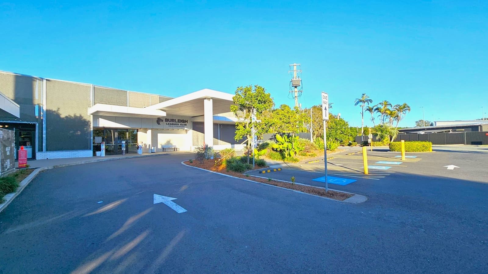 A building with a sign reading "Burleigh Leagues Club" is on the left, featuring a modern design with large windows and a flat roof. In front, there are landscaped areas with small trees and shrubs. The car park on the right has marked spaces, including accessible parking, and a "One Way" sign. The ground is paved, and there are yellow bollards near the parking spaces. In the background, there are more trees and a power pole.