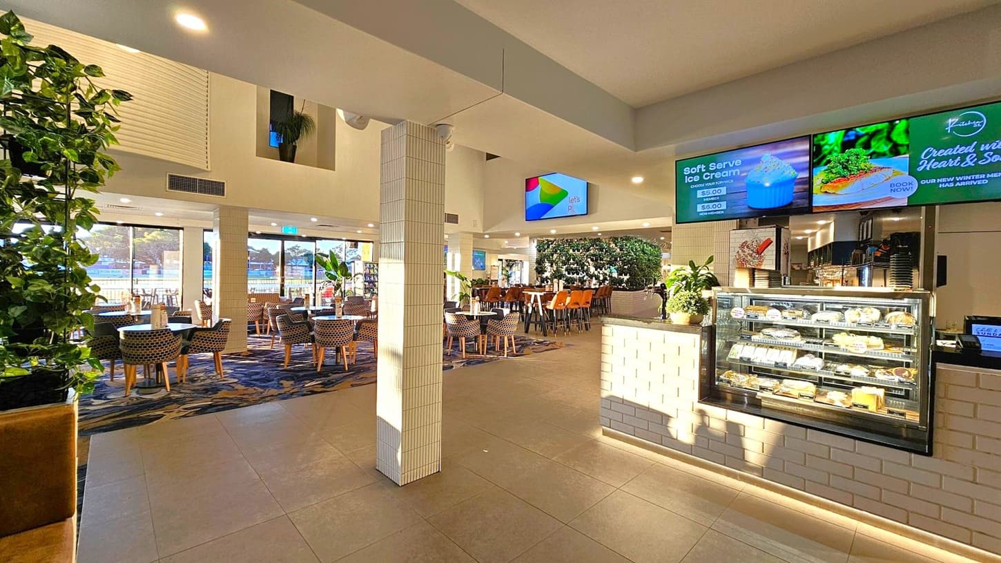 A spacious indoor dining area with a modern design. On the right, there's a counter with a glass display case filled with pastries and desserts. Above it, digital screens display menu items. In the centre, a tiled pillar divides the space. To the left, tables and chairs are arranged neatly, with large windows allowing natural light to flood in. The floor is tiled, and there are plants adding greenery throughout the space. The ceiling is high, with recessed lighting.