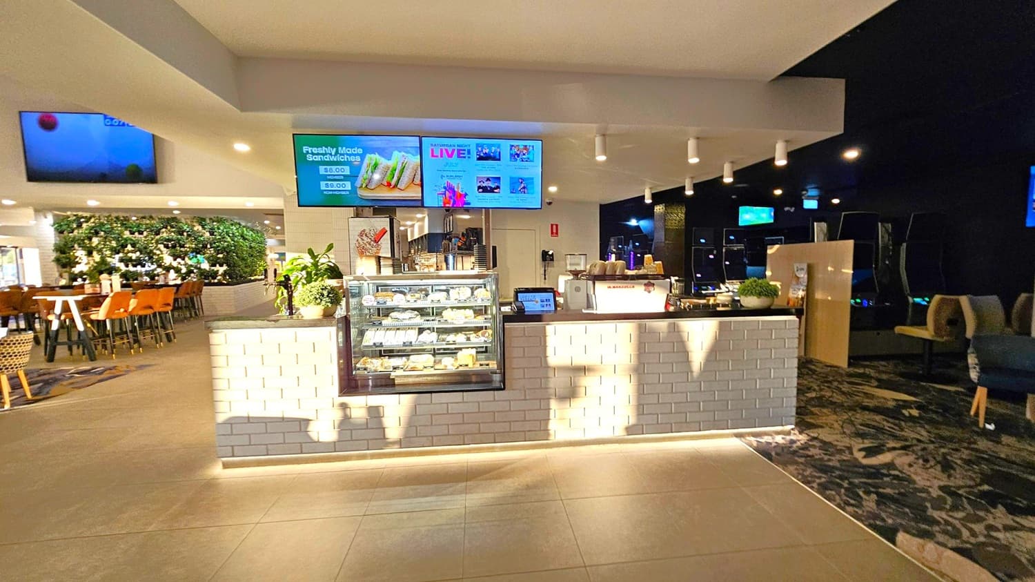A modern café counter with a white brick front. In the centre, a glass display case contains various sandwiches and pastries. Above, digital screens display menu items and promotions. To the left, a seating area with wooden tables and chairs is visible, along with a green plant wall. The right side is darker, with more seating and dim lighting. The floor is tiled, and the ceiling has recessed lighting.