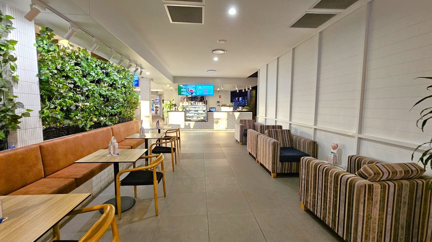 A modern café interior with a green wall on the left, featuring lush plants above a long brown cushioned bench. Wooden tables and chairs are arranged alongside. On the right, there are striped armchairs against a light-coloured panelled wall. The floor is tiled, and the ceiling has recessed lighting. In the background, a counter with a display case and digital menu screens is visible, with additional plants adding to the décor.