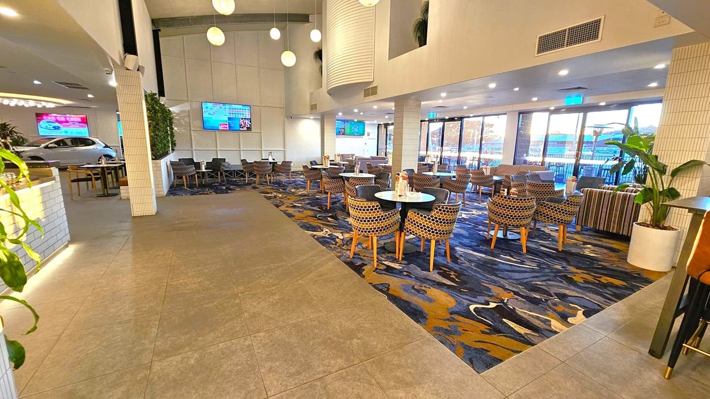 A spacious indoor dining area with a mix of tiled and patterned carpet flooring. On the left, there is a white-tiled counter and a car visible through a large window. In the centre, round tables with patterned chairs are arranged neatly. The right side features large windows with a view outside, and a potted plant is placed near the seating area. The ceiling is high with hanging spherical lights, and there are multiple screens mounted on the walls.