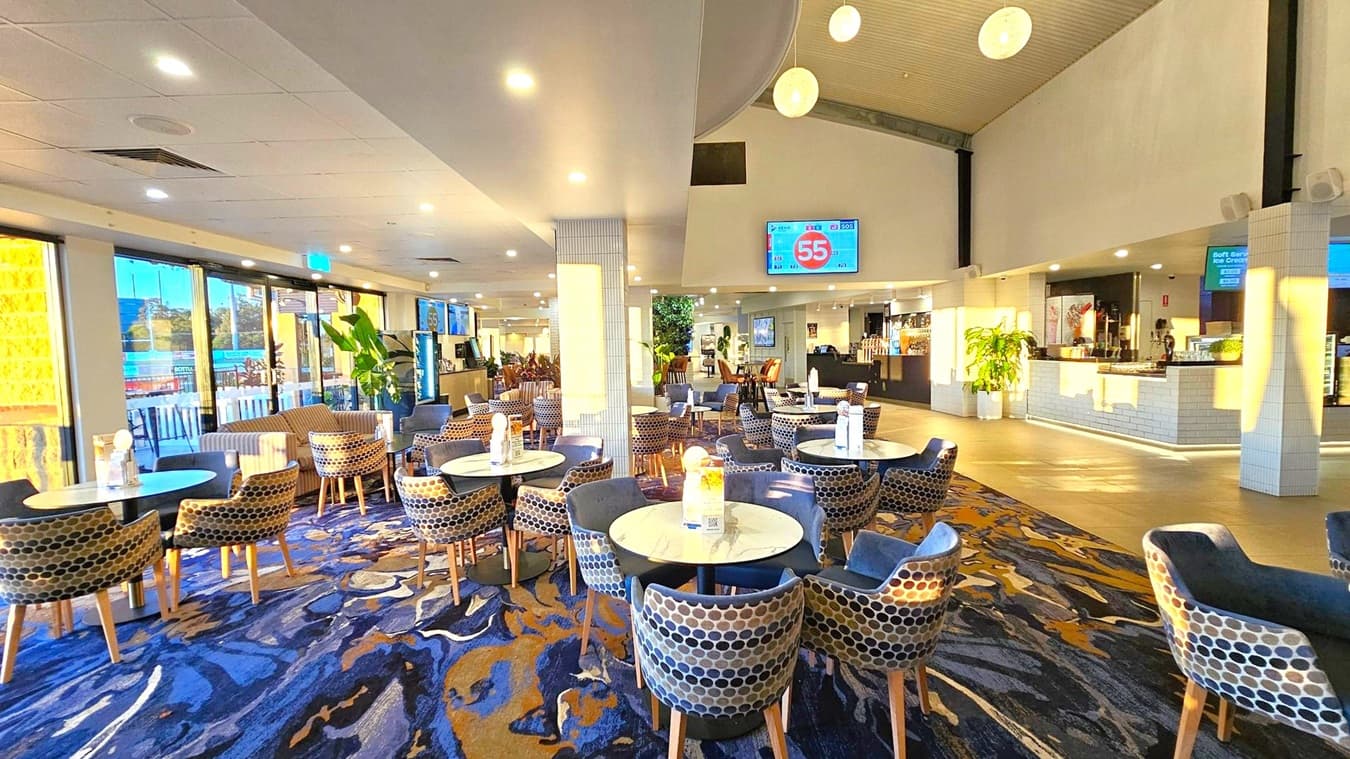 A spacious indoor dining area with round tables and patterned chairs arranged across a carpeted floor featuring a blue and gold abstract design. On the left, large windows allow natural light to illuminate the space, with a view of the outdoors. The right side has a counter with a menu display and potted plants. Overhead, spherical pendant lights hang from the ceiling, and a digital screen displays a number. The ceiling is white with recessed lighting.