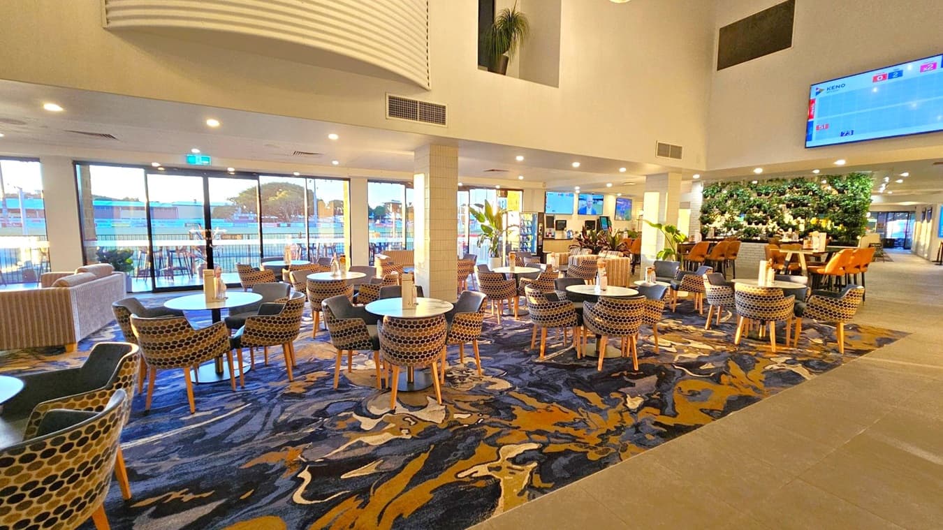 Spacious interior with a patterned carpet covering the floor. On the left, round tables with patterned chairs are arranged in rows. Large windows in the background allow natural light to flood in, revealing an outdoor view. On the right, a vertical garden wall adds greenery, with more seating and tables nearby. A large screen is mounted on the upper right wall. The ceiling is high with recessed lighting, and the overall atmosphere is modern and open.