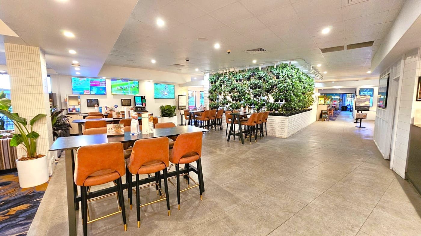 Spacious indoor dining area with a modern design. On the left, there are tables with brown chairs and digital screens displaying various content. A white-tiled column and potted plants add to the decor. In the centre, a green wall with lush plants separates sections of the room. The floor is tiled in grey, and the ceiling is white with recessed lighting. Towards the back, more seating is visible, along with a digital kiosk near the entrance.