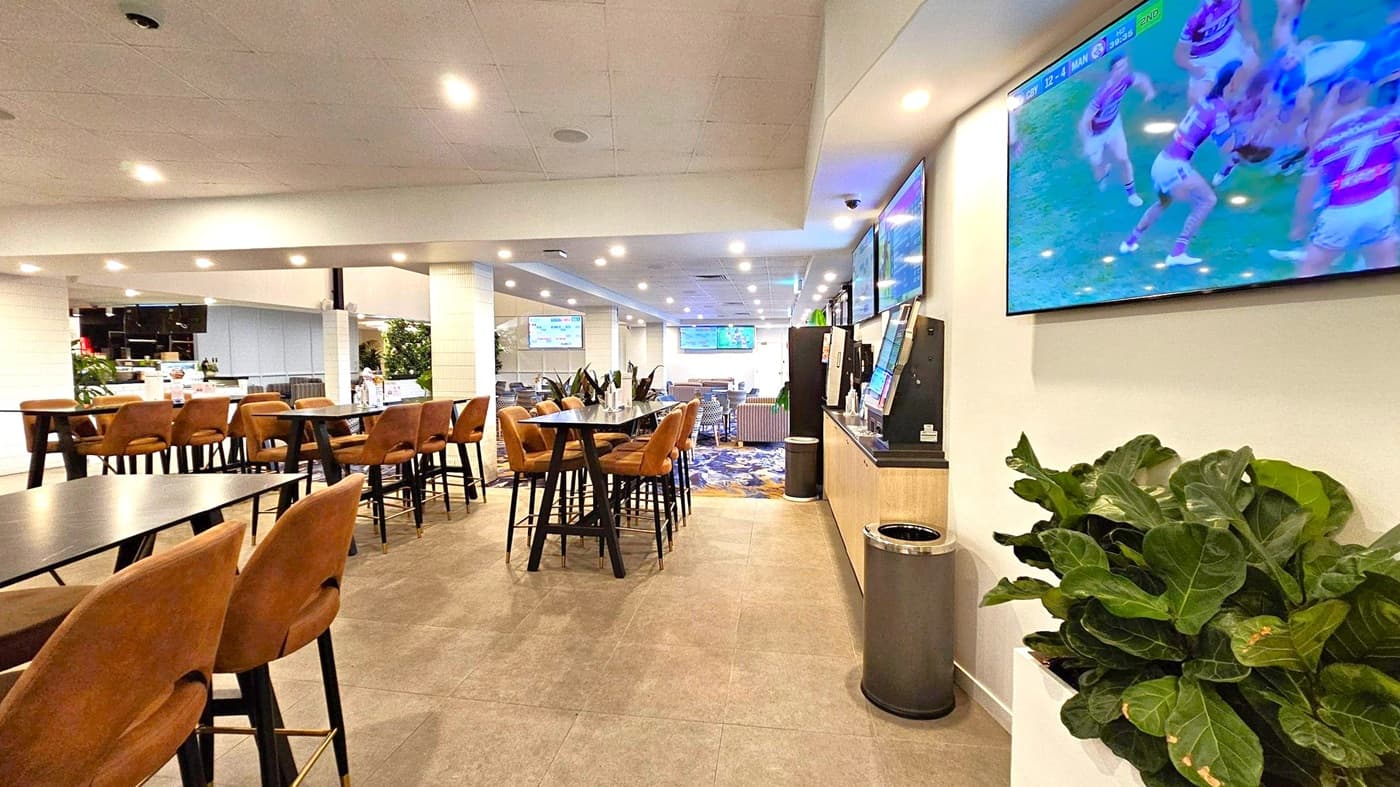 A modern indoor space with a high ceiling and recessed lighting. On the right, a large TV displays a sports game above a plant and a waste bin. In the centre, there are high tables with brown chairs, and a patterned carpet is visible further back. The left side features more seating and a counter area with additional plants. Multiple screens are mounted on the walls, and the floor is tiled in a neutral tone.