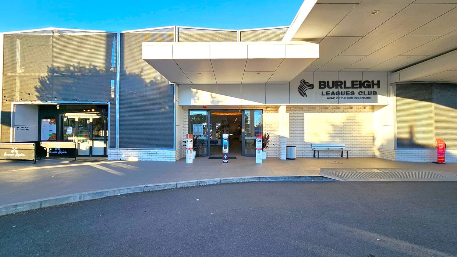 Entrance to a building with a sign reading "Burleigh Leagues Club" and "Home of the Burleigh Bears" on the right. The facade is made of light-coloured bricks. A bench and a bin are near the entrance. On the left, there's a smaller entrance with a sign for "Jasmine" and a canopy above. The ground is paved, and the ceiling has a modern, flat design. Shadows of trees are cast on the building.