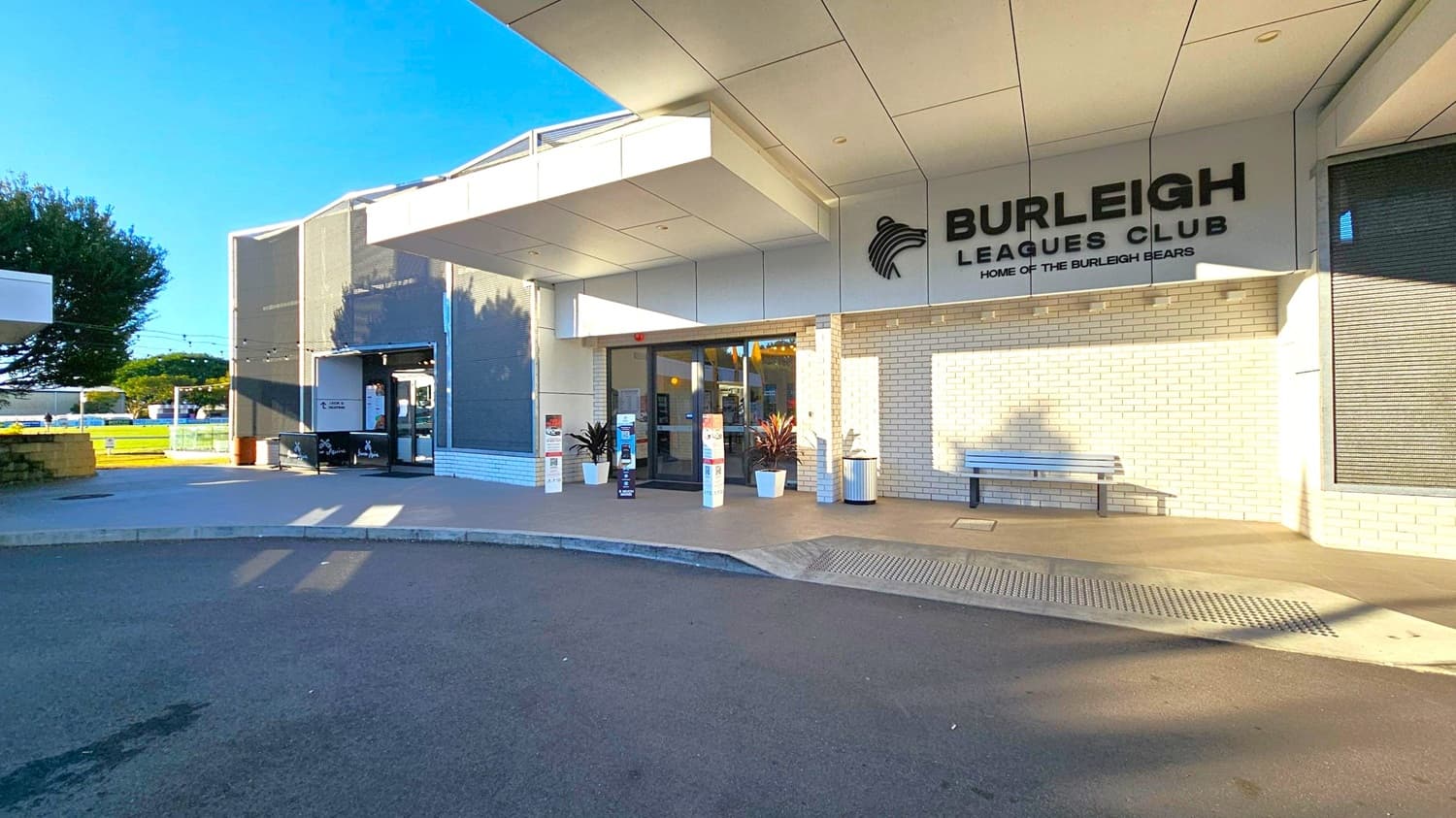 The image shows the entrance to the Burleigh Leagues Club. On the right, there's a sign with the club's name and logo on a white brick wall. A bench is positioned against the wall. In the centre, glass doors are flanked by potted plants and informational pillars. On the left, a tree and some outdoor seating are visible. The ceiling is modern with a white panel design, and the ground is paved with a mix of concrete and asphalt.