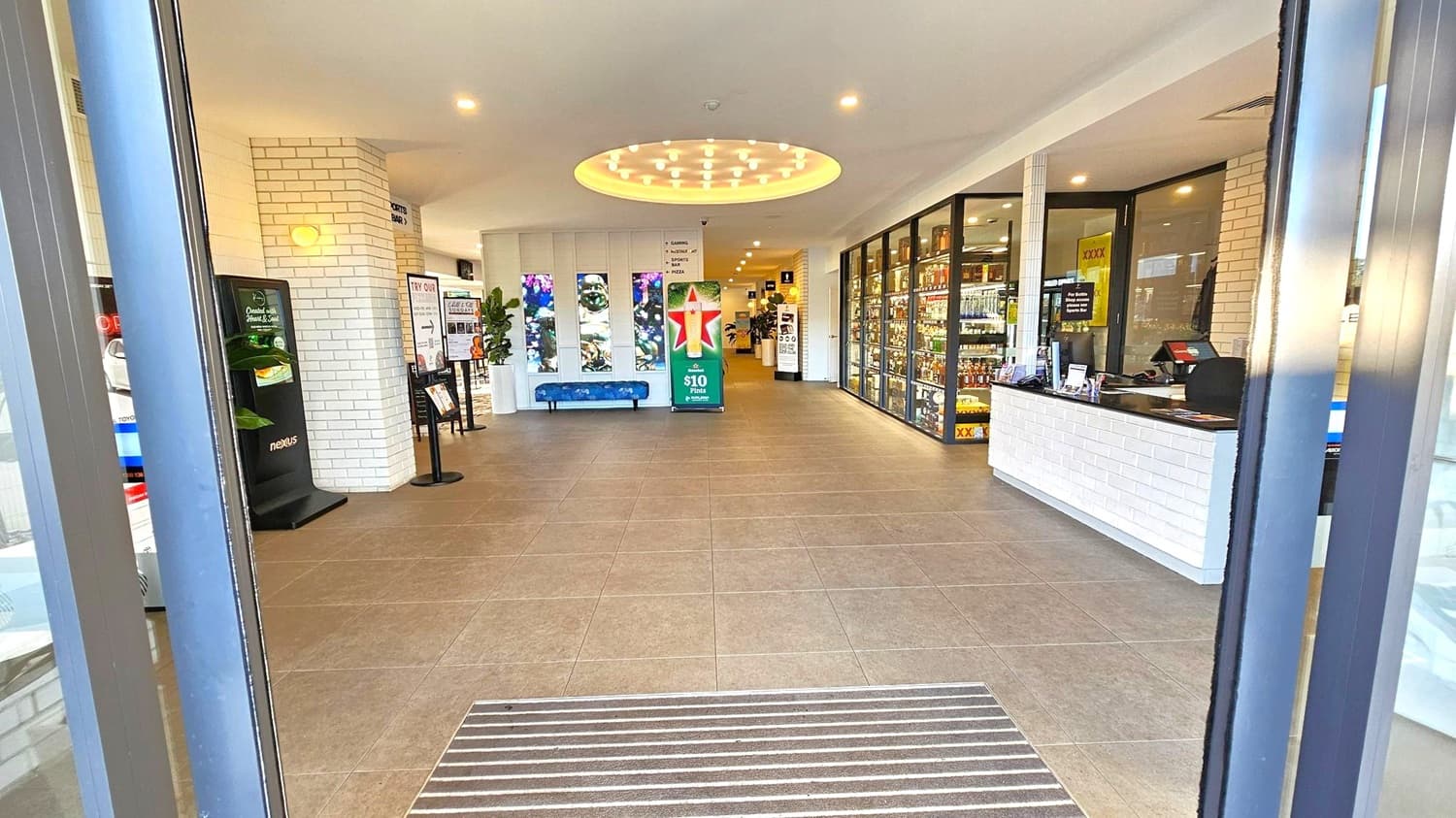 Entrance to a modern store with a tiled floor and a circular ceiling light fixture. On the left, a white brick wall with a digital display and signage. In the centre, a promotional stand and a blue bench against a wall with decorative panels. On the right, a counter with a cash register and a glass display of bottles. The space is well-lit with recessed lighting.
