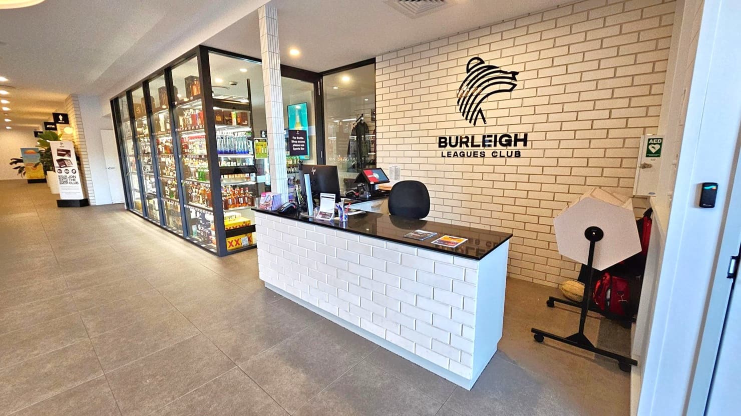 Reception area with a white brick counter topped with a black surface, featuring a computer and promotional materials. Behind, a white brick wall displays the "Burleigh Leagues Club" logo. To the left, a glass display case showcases various bottles. The floor is tiled in grey, and the ceiling has recessed lighting. A sign and a plant are visible further down the hallway.