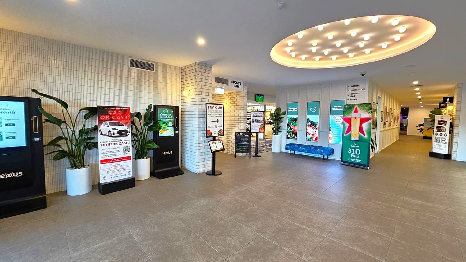 Spacious interior with a tiled floor and a ceiling featuring circular lighting. On the left, there are digital kiosks and promotional signs, flanked by large potted plants. In the centre, a column with more signage and a small menu stand. To the right, a hallway with illuminated displays and a blue bench. The walls are lined with white tiles and framed pictures. An ATM is visible in the background.