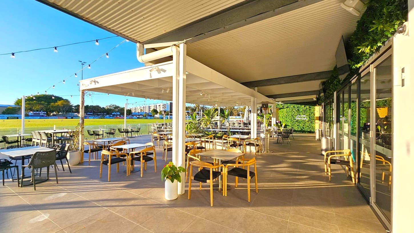 An outdoor dining area with a covered patio. On the left, there are tables and chairs arranged neatly, with string lights hanging above. The background features a grassy field and trees. On the right, glass doors reflect the patio, and there are more tables and chairs. The ceiling is corrugated, and potted plants are placed around the area, adding greenery. The floor is tiled, and the space is well-lit by natural light.