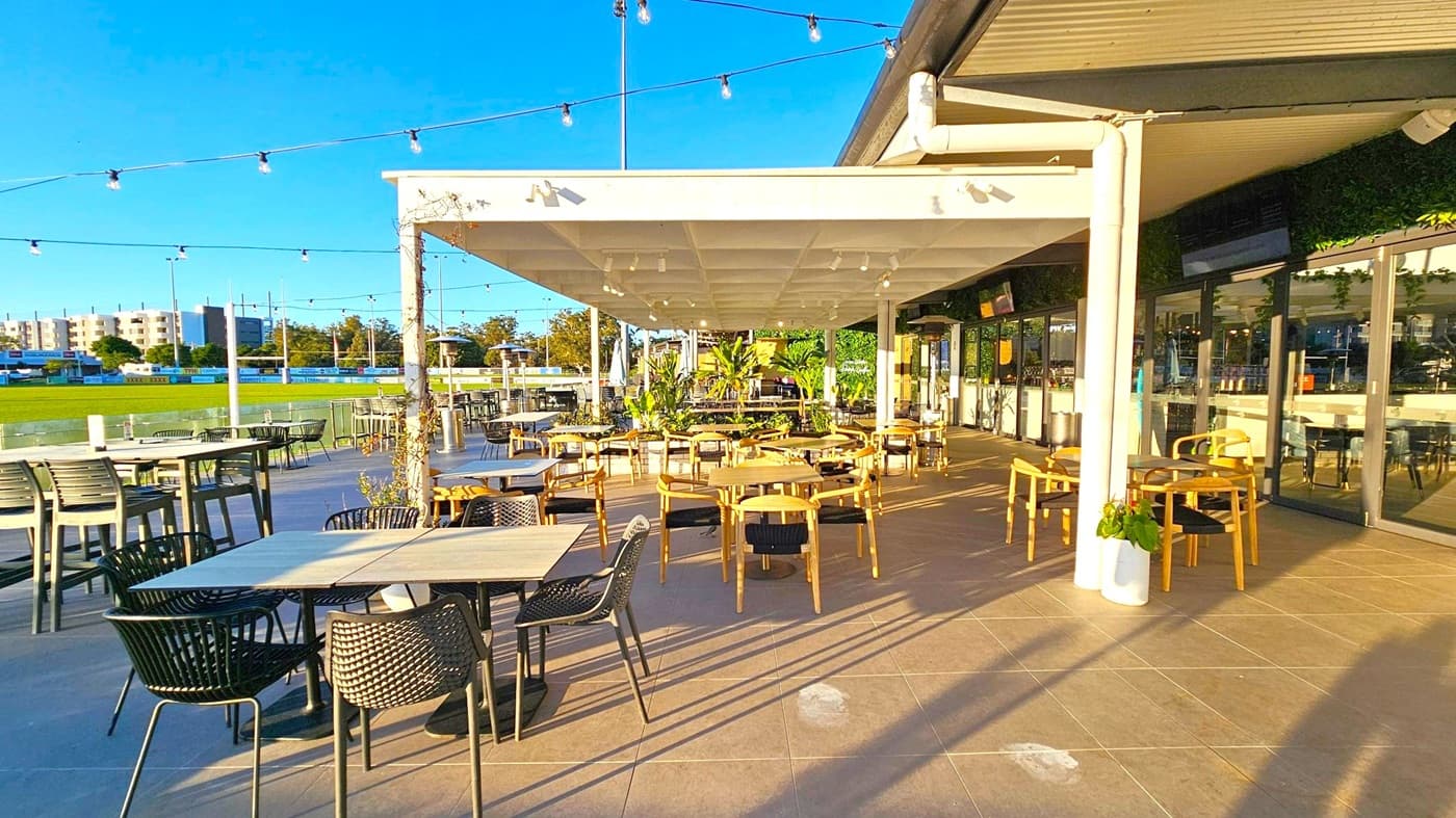 Outdoor dining area with a mix of tables and chairs, some under a white pergola. String lights are suspended above. On the left, a grassy field and buildings are visible in the background. The right side features glass doors leading inside. The floor is tiled, and there are potted plants around the seating area.