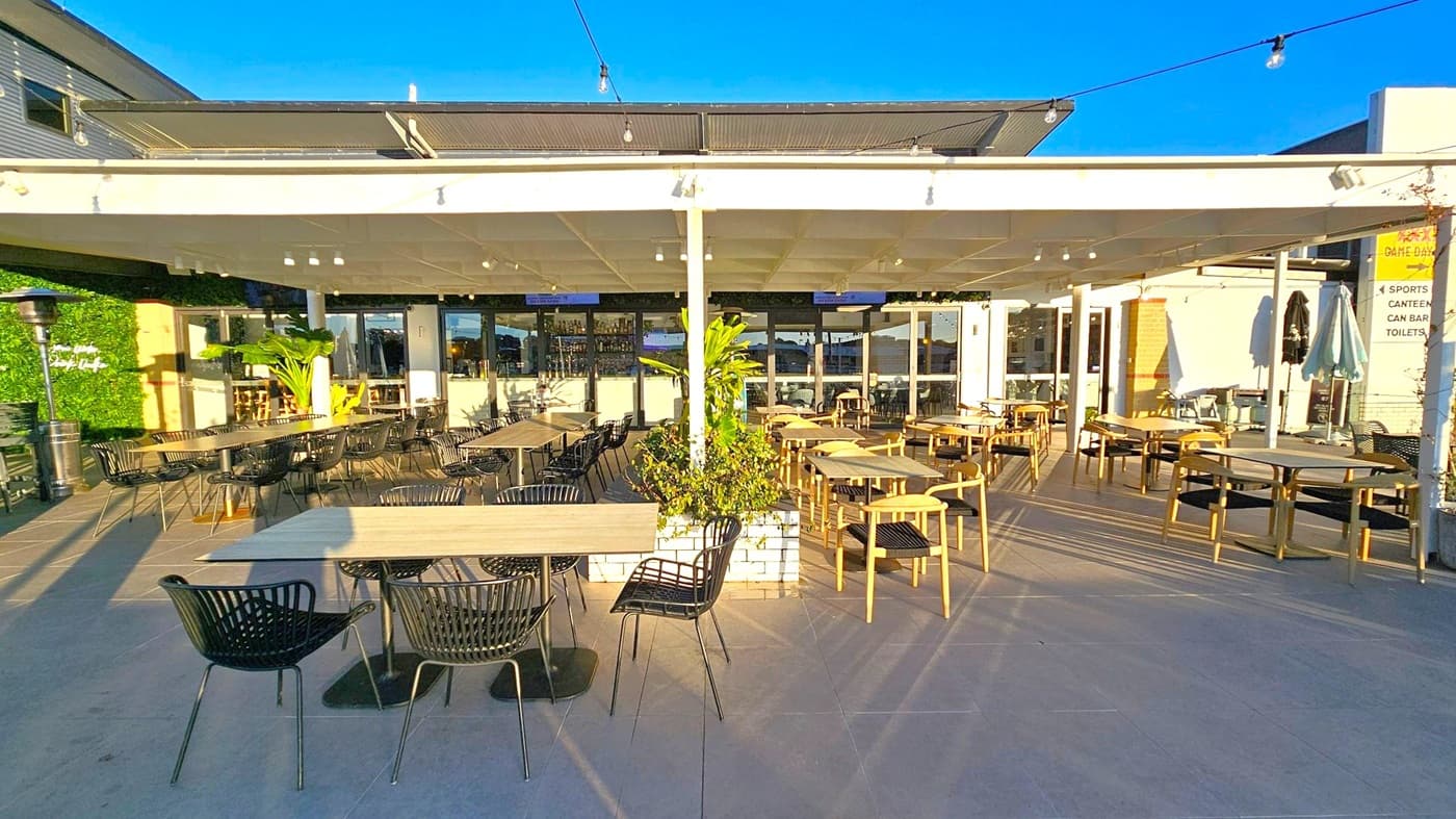 Outdoor seating area with a mix of black and wooden chairs around rectangular and round tables. A white pergola with string lights covers the space. In the centre, a planter with green plants adds a touch of nature. The background features a building with large windows and a bar area. The floor is tiled, and there are patio heaters and umbrellas scattered around. A sign on the right indicates directions to various facilities.
