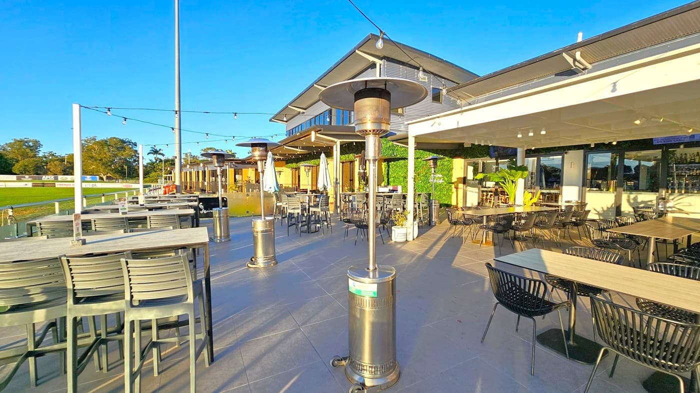 Outdoor dining area with several tables and chairs on a tiled floor. Patio heaters are spaced throughout the area. On the left, there are high tables with bar stools, and a grassy field with advertising boards in the background. The right side features standard tables with black chairs under a white pergola. The building in the background has large windows and a sloped roof. String lights are hung above the seating area.