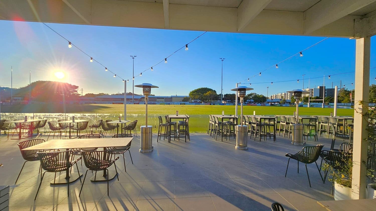 Outdoor seating area with tables and chairs on a tiled floor. String lights are hung above, and there are tall patio heaters. In the background, a grassy field is visible with goalposts and a few trees. The sun is low in the sky, casting long shadows across the area. Buildings are visible in the distance. The ceiling is covered with beams.
