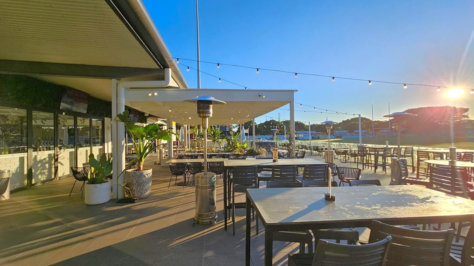 Outdoor dining area with several tables and chairs on a patio. On the left, a building with large windows and potted plants. Overhead, string lights are suspended across the space. In the centre, a patio heater stands among the tables. The background features a grassy area with trees and a bright sun low in the sky, casting long shadows across the ground. The floor is tiled, and the ceiling of the covered section is white.