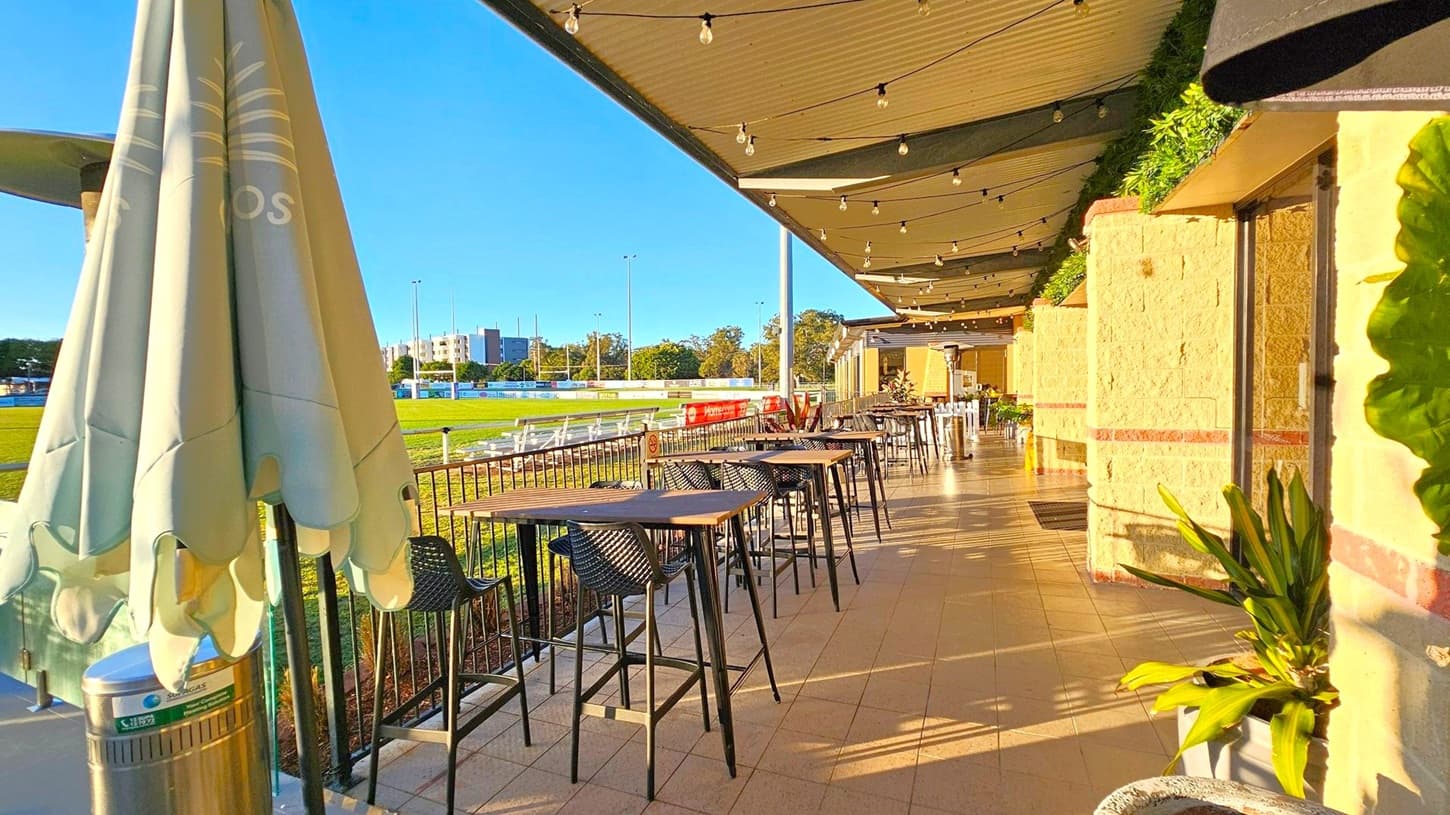 A covered outdoor seating area with a row of high tables and chairs on the right. The floor is tiled, and the ceiling has string lights. On the left, there's a closed umbrella next to a metal bin. Beyond the railing, a grassy field is visible with some buildings in the distance. The wall on the right is light-coloured with a red stripe, and there are potted plants along the walkway.