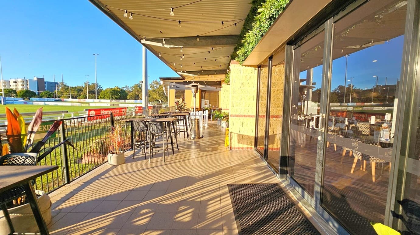 A covered outdoor seating area with a tiled floor and string lights hanging from the ceiling. On the right, there are large glass doors reflecting the surroundings, leading into an interior space with tables and chairs. On the left, a black metal railing separates the area from a grassy field with a racetrack and advertising boards. Several high tables with black woven chairs are arranged along the railing, and potted plants are placed intermittently.