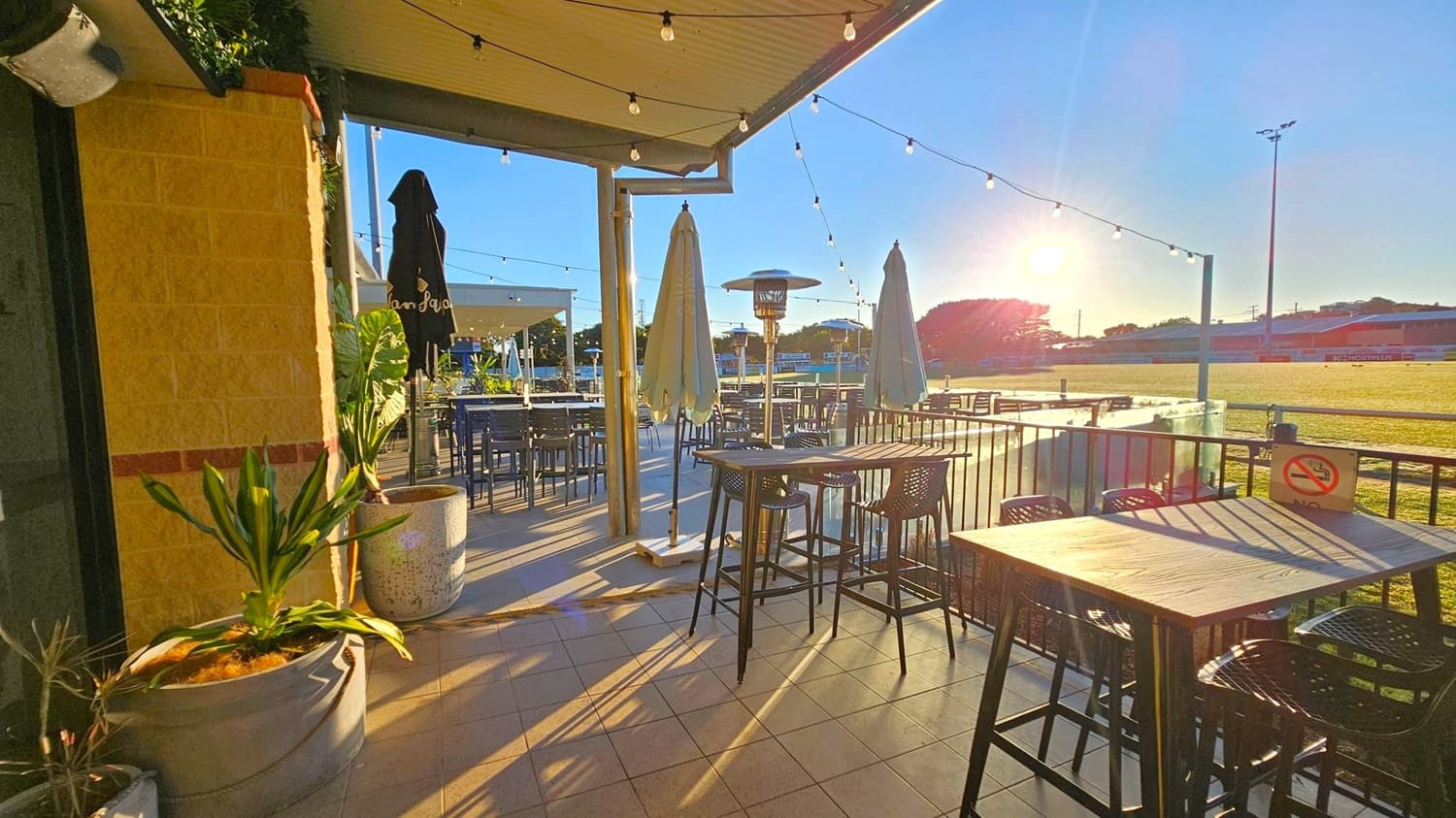 Outdoor seating area with several tables and high chairs on a tiled floor. On the left, a brick wall with potted plants. In the centre, two closed umbrellas and a patio heater. String lights hang from the ceiling. The background features a grassy field with a tall light pole and a low building. The sun is low in the sky, casting long shadows across the area. A "No Smoking" sign is visible on one of the tables.