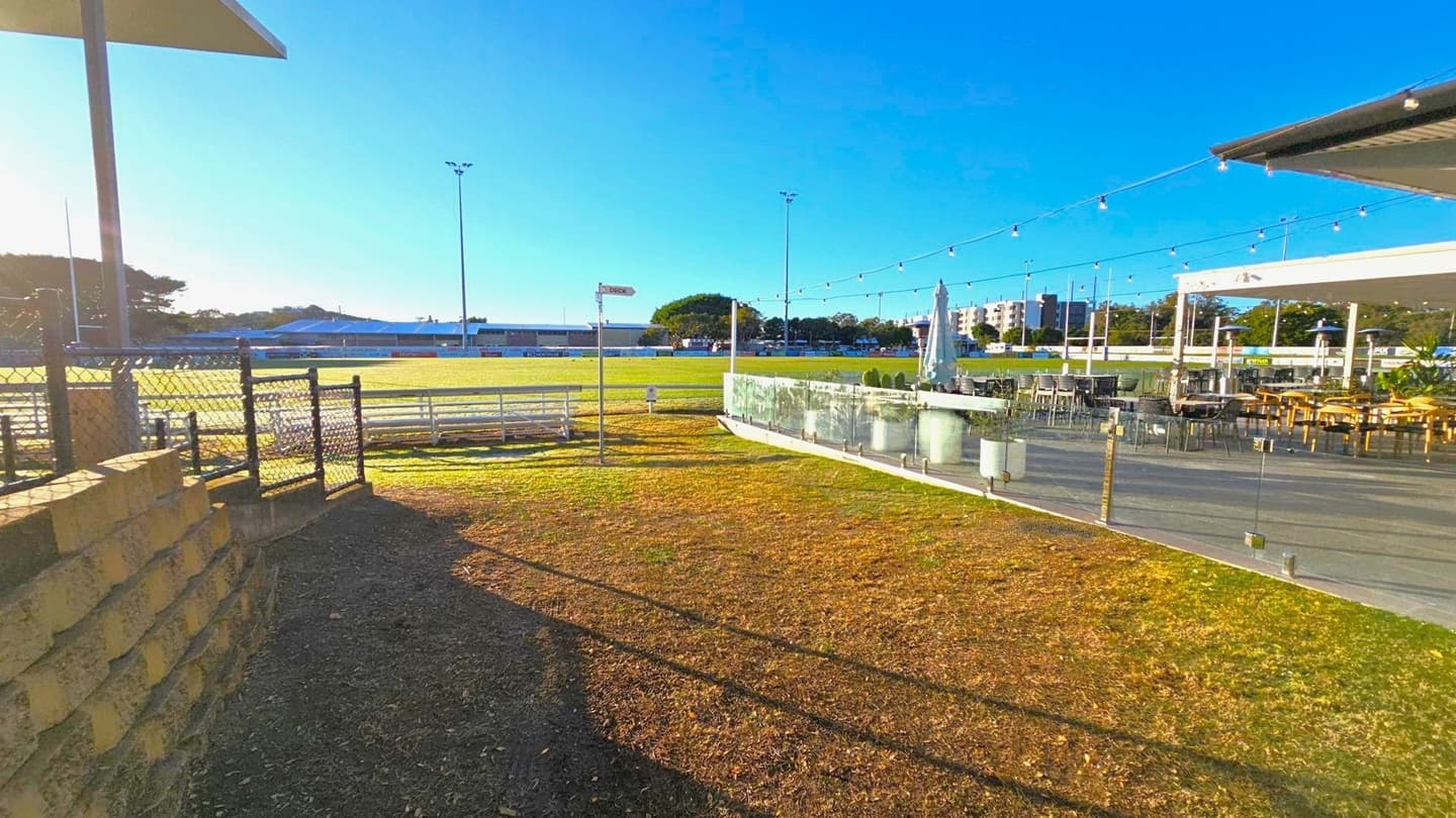A grassy area with a chain-link fence and a stone wall on the left. In the centre, a sports field is visible with tall floodlights and a signpost. On the right, there is a patio area with tables, chairs, and an umbrella, enclosed by a glass barrier. String lights are hung above the patio. In the background, there are buildings and trees. The ground is a mix of grass and dirt, and the sky is clear.