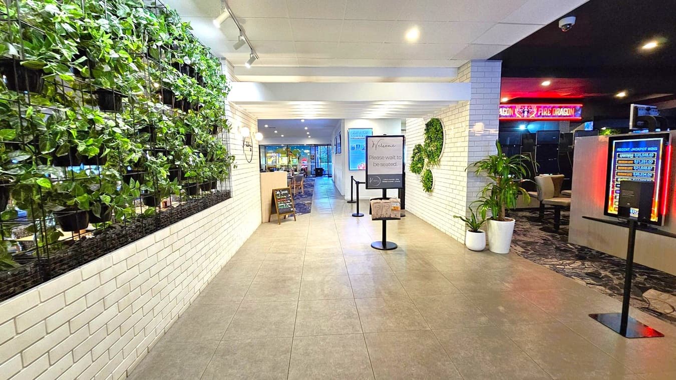 A corridor with a tiled floor and a white ceiling. On the left, a wall features a vertical garden with lush green plants. The right side has white brick walls with circular plant displays and a digital sign showing recent jackpot wins. In the centre, a sign reads "Welcome, Please wait to be seated," with a menu stand nearby. In the background, a restaurant area is visible, and to the right, a section with a "Fire Dragon" sign is partially visible.