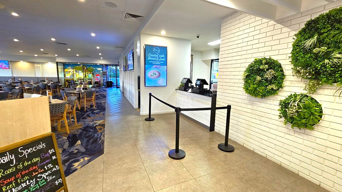 A restaurant interior with a tiled floor and a white ceiling with recessed lighting. On the right, a white brick wall features circular green plant decorations. A counter with a digital display is near the entrance, separated by a black rope barrier. On the left, a dining area with tables and chairs is visible, with a menu board displaying daily specials. Large windows in the background show an outdoor view.