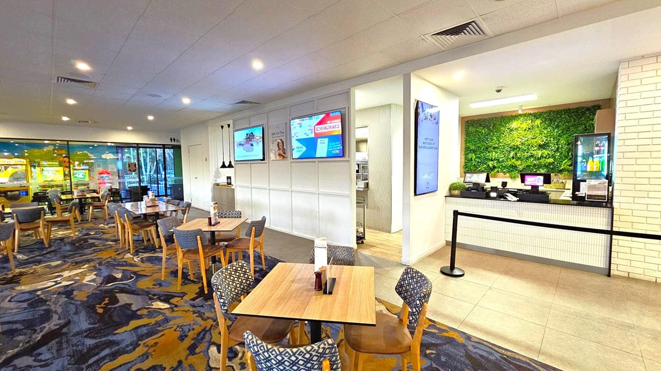 A modern indoor dining area with wooden tables and cushioned chairs on a patterned carpet. On the left, there are several tables and chairs, with a glass wall showing arcade machines outside. In the centre, three digital screens are mounted on a white wall. On the right, a counter with a green plant wall behind it and a small fridge displaying drinks. The ceiling is white with recessed lighting, and the floor near the counter is tiled.