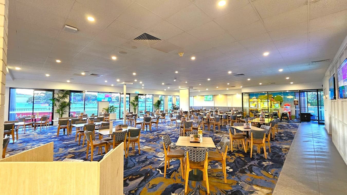 A spacious dining area with numerous wooden tables and chairs arranged neatly across a patterned carpeted floor. Large windows on the left and right walls allow natural light to enter, with views of an outdoor area. The ceiling is fitted with recessed lighting, and there are several potted plants near the windows. A few television screens are mounted on the walls, displaying various content. The overall atmosphere is open and inviting.