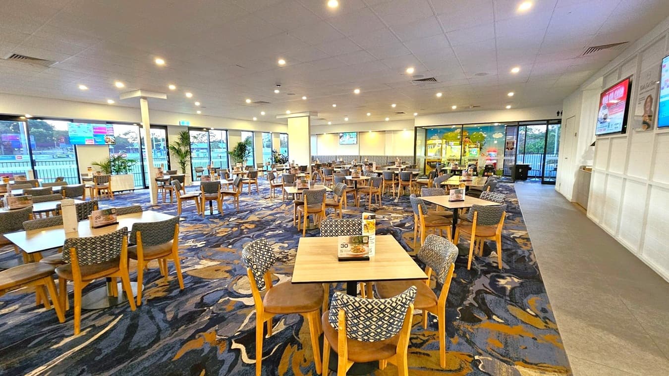 A spacious dining area with numerous wooden tables and chairs, each set with menus. The floor is covered with a patterned carpet in shades of blue and brown. Large windows on the left provide a view of the outdoors, while the right side features a wall with mounted screens displaying advertisements. The ceiling is fitted with recessed lighting, and there are potted plants near the windows. In the background, a section with arcade games is visible.