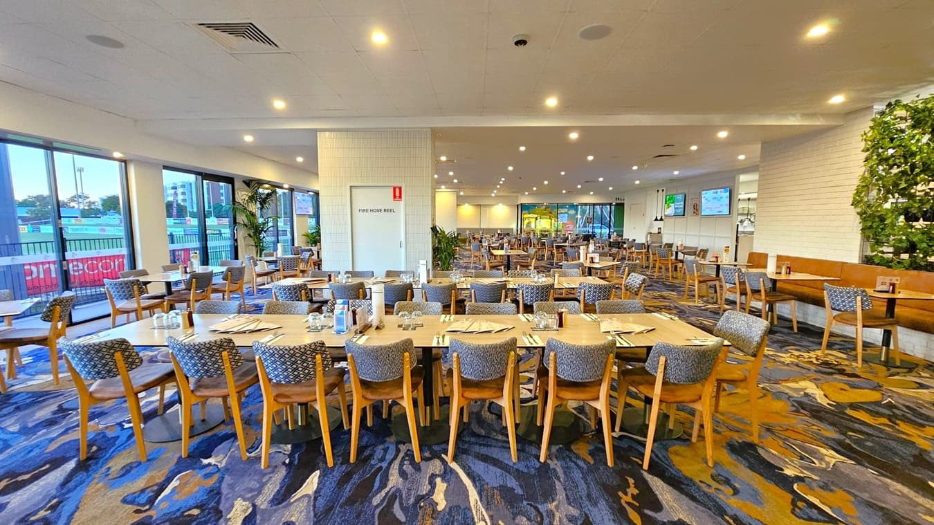 A spacious dining area with numerous wooden tables and chairs arranged neatly. The floor is covered with a patterned carpet in shades of blue and brown. On the left, large windows provide a view of an outdoor area. In the centre, a door marked "Fire Hose Reel" is visible. The right side features a wall with a vertical plant display. The ceiling is fitted with recessed lighting, creating a well-lit environment.