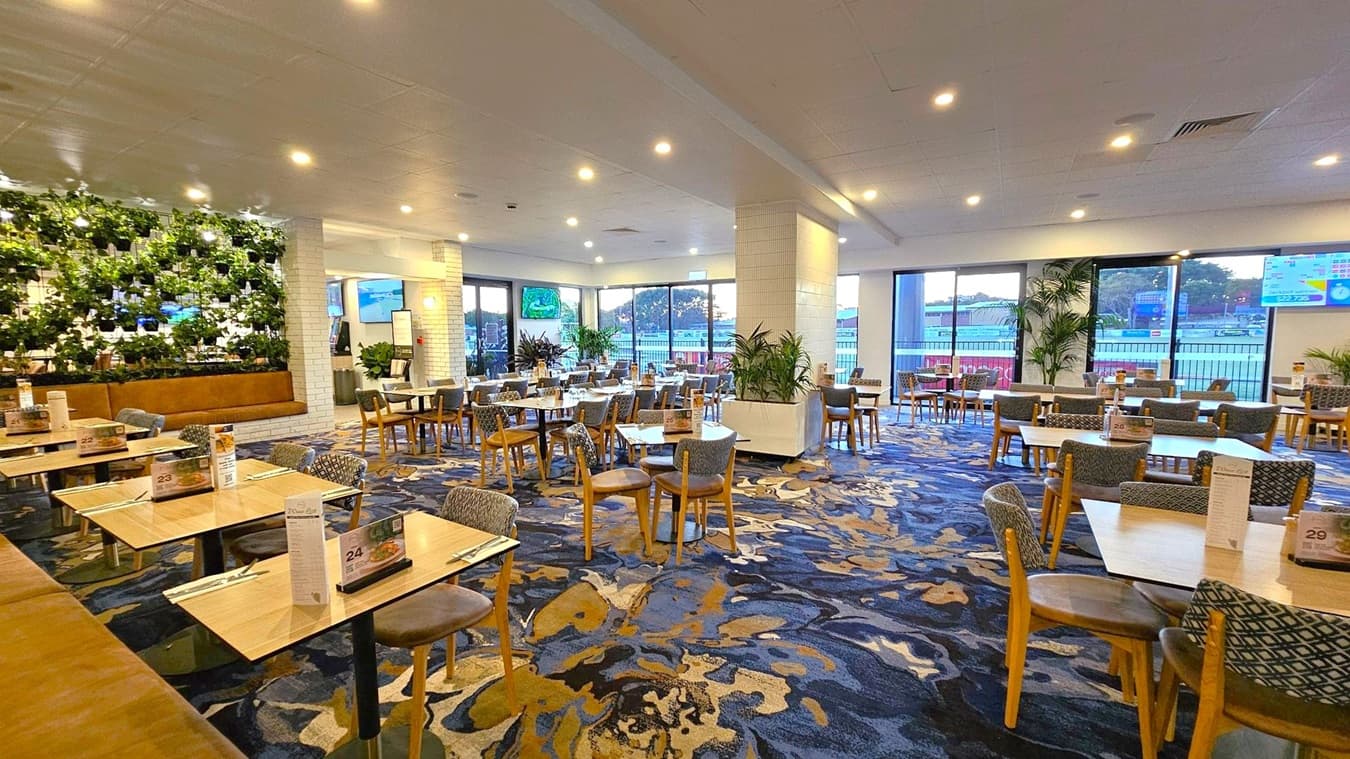 A spacious dining area with a patterned carpet in shades of blue and brown. On the left, there's a wall with green plants and a brown cushioned bench. Several wooden tables and chairs are arranged throughout the room, each with menus and cutlery. Large windows on the right let in natural light, and there are potted plants near the windows. The ceiling is white with recessed lighting, and there are TV screens mounted on the walls.