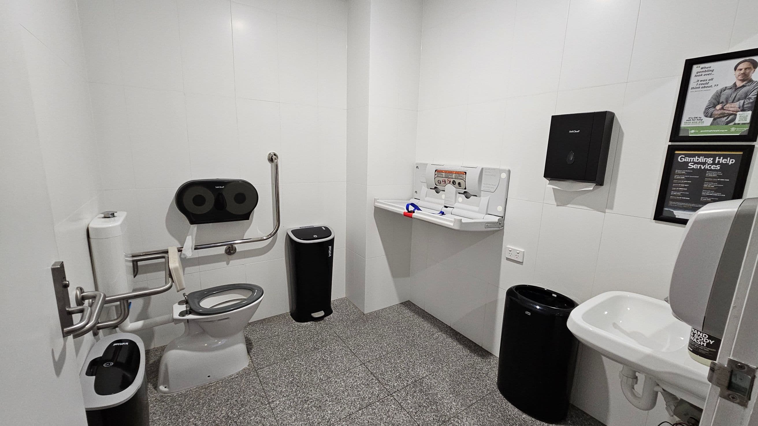 A public restroom with a grey tiled floor and white walls. On the left, there is a toilet with a support rail and a dual toilet paper dispenser above it. A sanitary bin is positioned nearby. In the middle, a baby changing station is mounted on the wall. On the right, a sink with a soap dispenser is visible, along with a black paper towel dispenser above a bin. Two framed posters are on the wall, one about gambling help services. The ceiling is plain white with recessed lighting.
