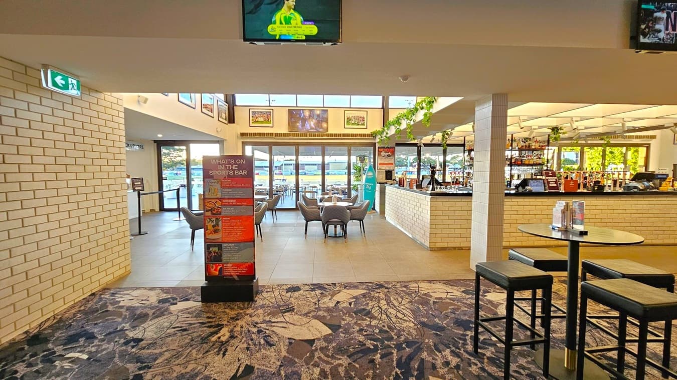 A sports bar interior with a patterned carpeted floor. On the left, a white brick wall with an exit sign. In the centre, a signboard listing events and a seating area with tables and chairs near large windows. The right side features a bar with a white brick counter, shelves of bottles, and a high table with stools. The ceiling has a grid pattern with hanging plants. Televisions are mounted above, displaying sports content.