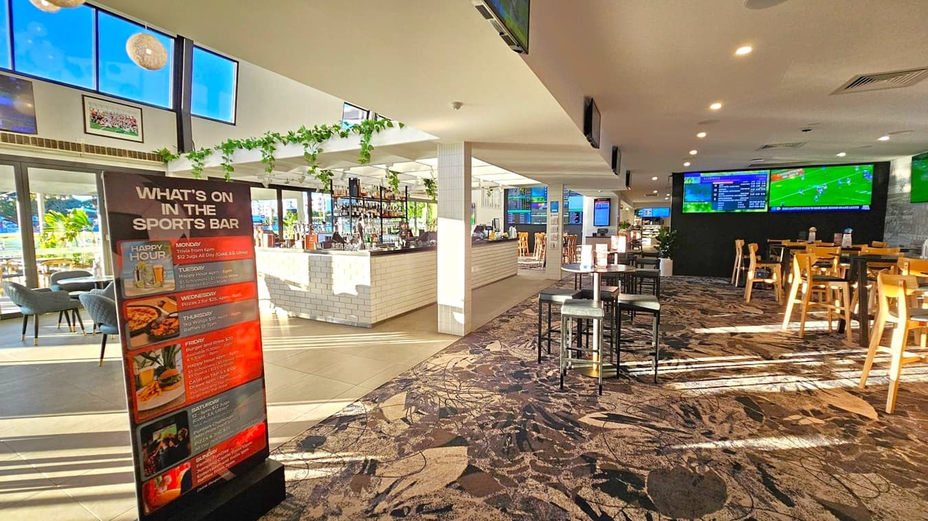 A sports bar interior with a large sign on the left listing weekly events. The bar area in the centre features a white brick counter adorned with hanging plants. On the right, there are tables and chairs on a patterned carpet. Multiple screens display sports content on the walls. The ceiling is white with recessed lighting, and the floor transitions from tiled to carpeted. Large windows in the background allow natural light to illuminate the space.