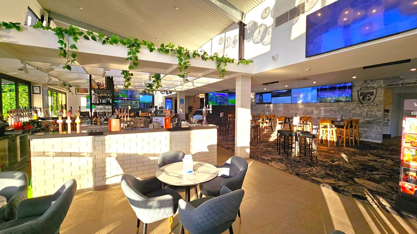 A modern bar interior with a central bar counter made of light-coloured bricks. The bar is equipped with multiple beer taps and overhead shelves holding bottles. Green plants hang from the ceiling beams. On the left, there are large windows allowing natural light in. In the foreground, a round table with four dark upholstered chairs. The background features several high tables and chairs, with multiple TV screens mounted on the walls. The floor is a mix of tiles and patterned carpet.