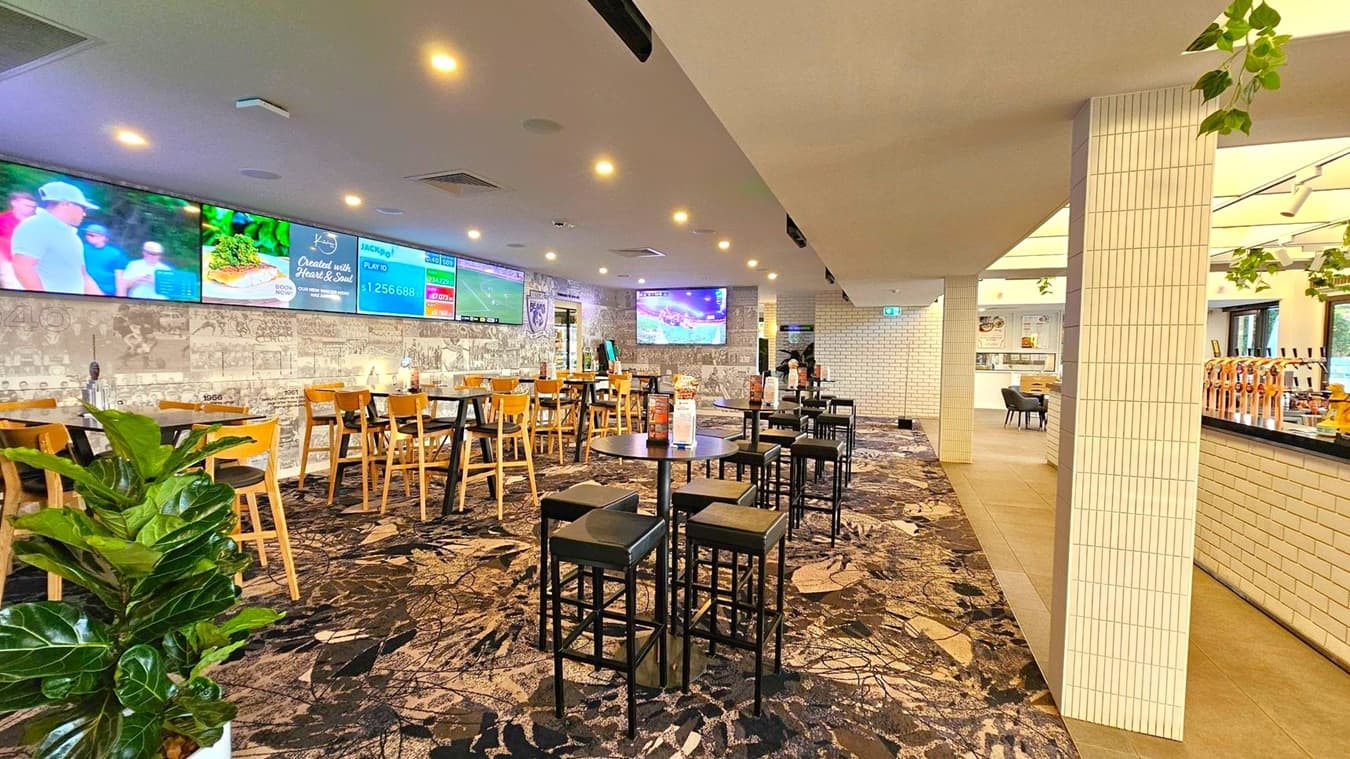 A spacious indoor area with a patterned carpet floor. On the left, there are wooden tables and chairs, with a large plant in the foreground. The wall features multiple screens displaying various images and information. In the middle, there are high tables with stools. On the right, a bar area with taps and a tiled wall. The ceiling has recessed lighting and some hanging plants. The background shows a white brick wall with an exit sign.