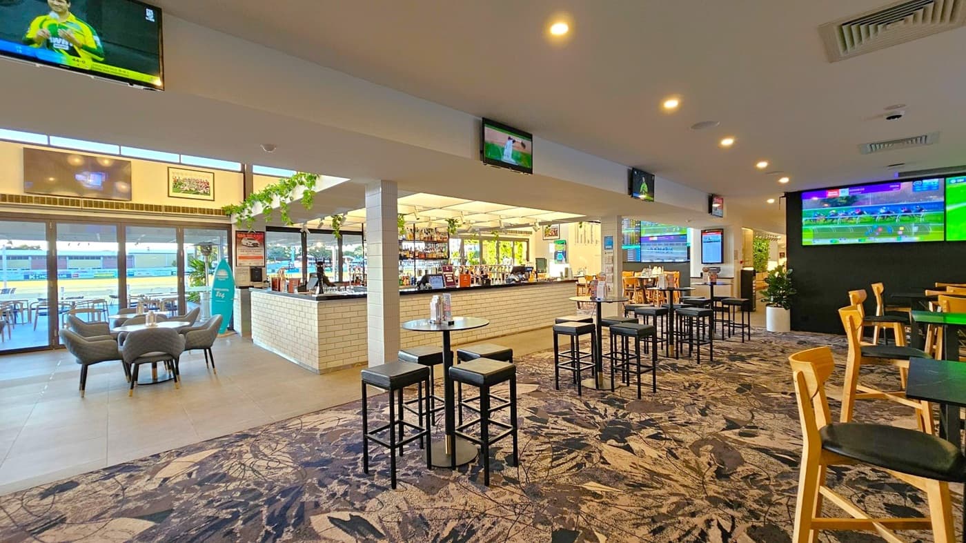 A spacious bar area with a patterned carpet floor. On the left, there are glass doors leading to an outdoor seating area with tables and chairs. Inside, there are several high tables with stools and a few armchairs around a low table. The bar counter is in the centre, with a variety of bottles displayed. Multiple screens are mounted on the walls, showing sports events. The ceiling is fitted with recessed lighting, and there are decorative plants hanging near the bar.