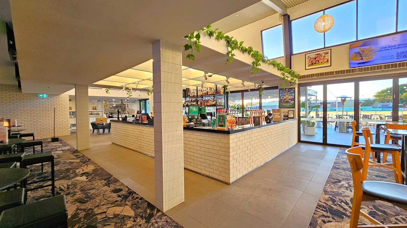 A modern bar area with a central counter made of light-coloured bricks and a black countertop. Overhead, green vines hang from the ceiling. To the left, there are tables and chairs on a patterned carpet. The right side features large windows and glass doors leading to an outdoor seating area with tables and chairs. The floor is a mix of tiles and carpet, and the ceiling is light-coloured with a hanging light fixture. Posters and signs are visible on the walls.