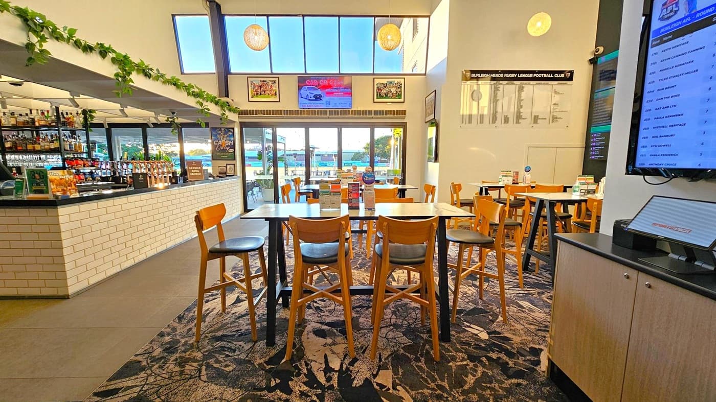 A spacious interior with a bar on the left featuring a white brick counter and shelves stocked with bottles. Greenery decorates the top of the bar. In the centre, there are high tables and wooden chairs on a patterned carpet. Large windows in the background let in natural light, showing an outdoor area. On the right, a wall-mounted screen displays information, and a cabinet is below it. The ceiling is high with hanging lights.