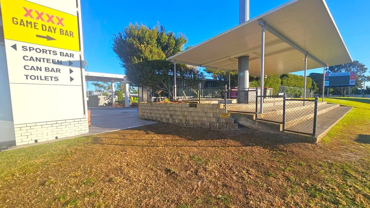 A sign on the left reads "XXXX Game Day Bar" with arrows pointing to various facilities like the sports bar, canteen, can bar, and toilets. To the right, there's a covered seating area with a metal fence and a small set of steps. Behind it, trees and shrubs provide greenery. In the background, a large scoreboard is visible on a grassy area. The ground is a mix of grass and dirt, and the structure has a flat roof supported by metal poles.