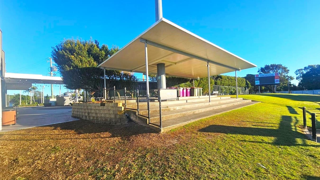A covered outdoor area with a large, flat roof supported by metal poles. Beneath the roof, there are several purple bins and a sign. The ground is a mix of grass and concrete steps leading up to the covered area. To the left, there is a building with a flat roof and some trees. In the background, a sign with the word "INSURANCE" is visible, along with more trees and a clear blue sky.