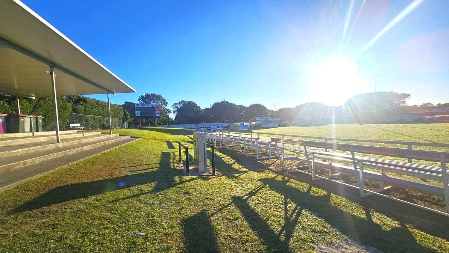 On the left, there is a covered seating area with several rows of concrete steps and a few bins. In the middle, a grassy field extends towards the right, with metal bleachers casting long shadows on the grass. In the background, a scoreboard is visible among trees, and the sun is shining brightly, creating lens flare. The sky is clear and blue.