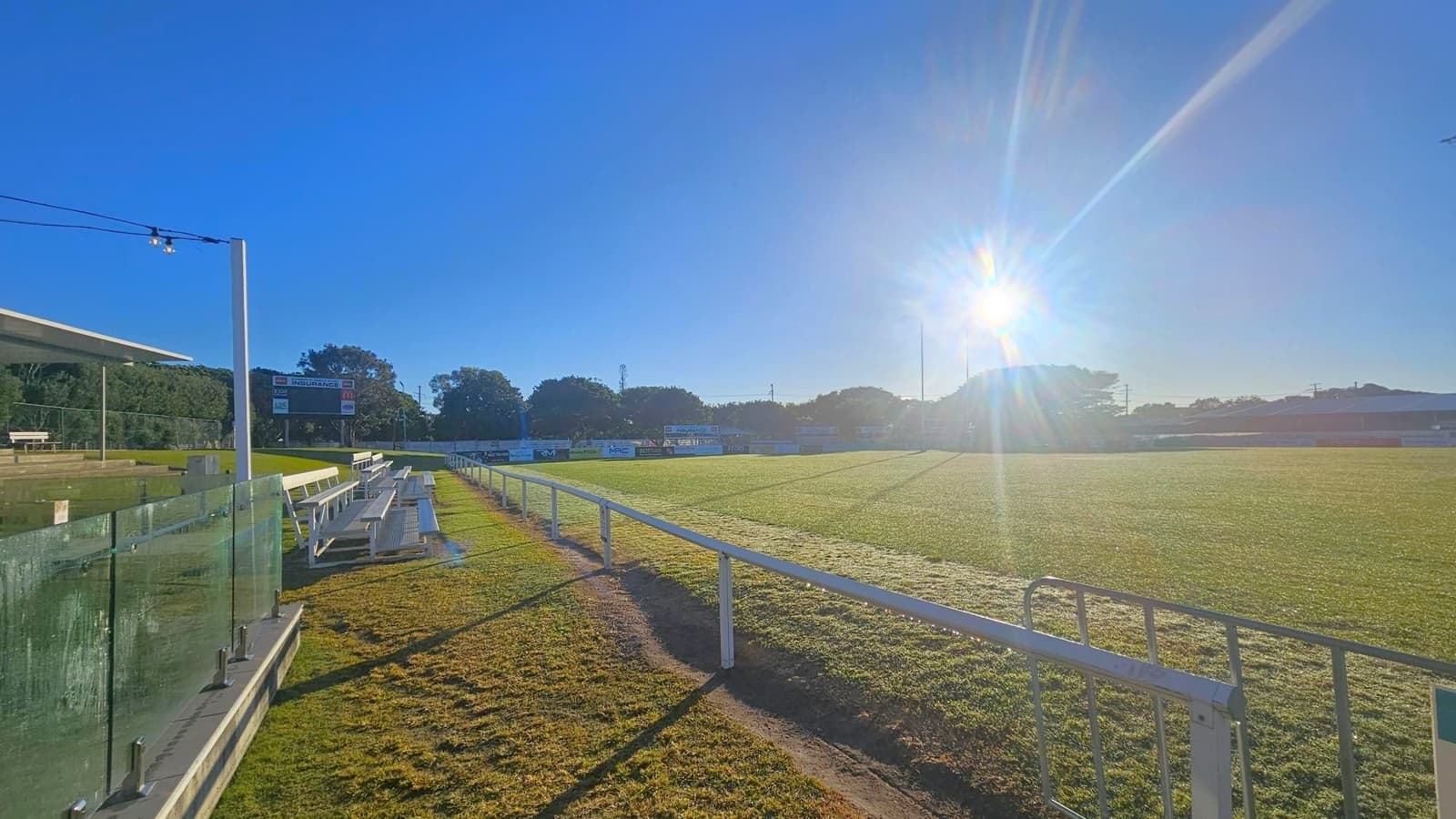 A sports field with a grassy surface is shown, with a bright sun low in the sky casting long shadows. On the left, there are tiered metal bleachers with a glass barrier in front. A scoreboard is visible in the distance, surrounded by trees. A white railing runs along the edge of the field, and the ground is slightly uneven with patches of grass. The sky is clear and blue.